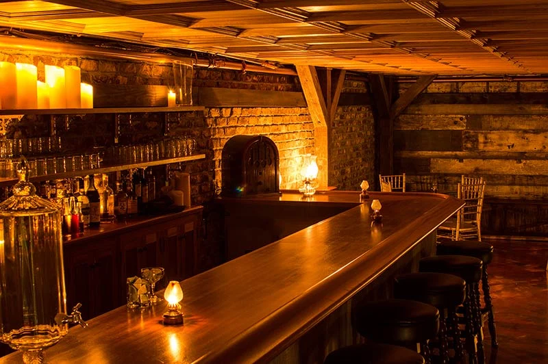 A long wooden bar beneath an ornate ceiling, with lamps, bottles, and barstools